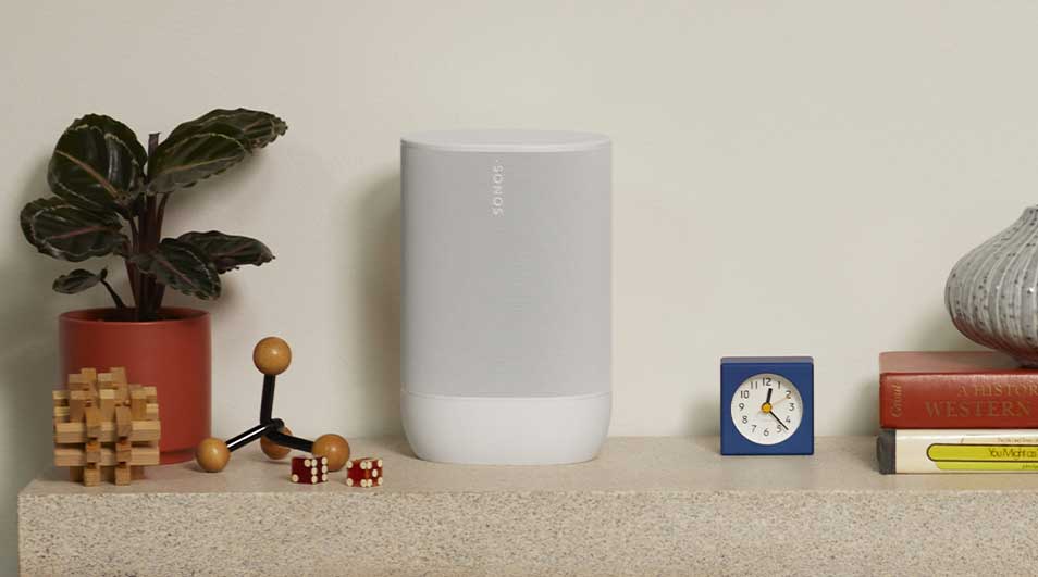 Een Sonos speaker staat op een plank, samen met een plant in een oranje pot, een blauwe klok, boeken, en decoratieve objecten.