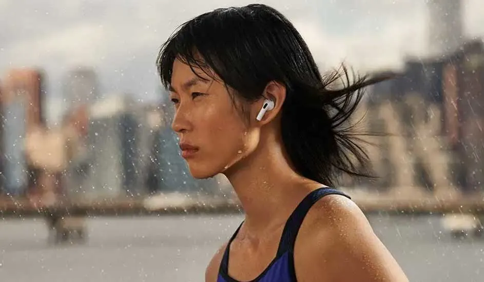 Une femme portant des oreillettes blanches est à l'extérieur sous la pluie, avec des bâtiments flous en arrière-plan.