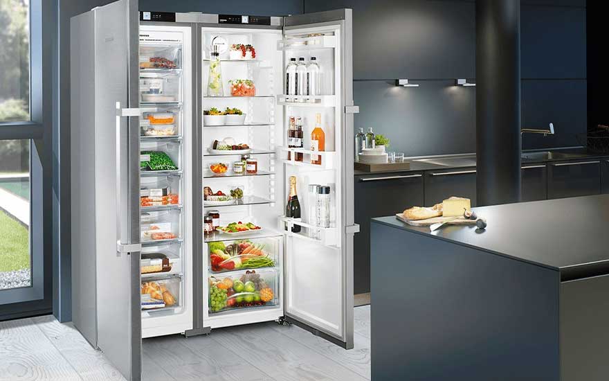 Een openstaande, zilverkleurige koelkast toont gevulde schappen met eten in een moderne keuken met donkere kasten en een eiland.