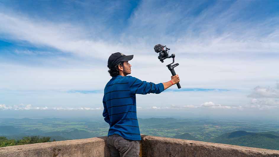 Sur une terrasse, une caméra noire avec microphone est tenue à bout de bras. Au loin, paysage de collines vertes sous ciel bleu nuageux.