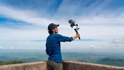 Sur une terrasse, une caméra noire avec microphone est tenue à bout de bras. Au loin, paysage de collines vertes sous ciel bleu nuageux.