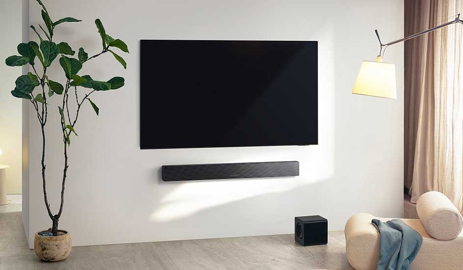Un téléviseur Samsung est fixé au mur, avec une barre de son Samsung en dessous et un caisson de basses dans un salon moderne avec plante et fauteuil design.