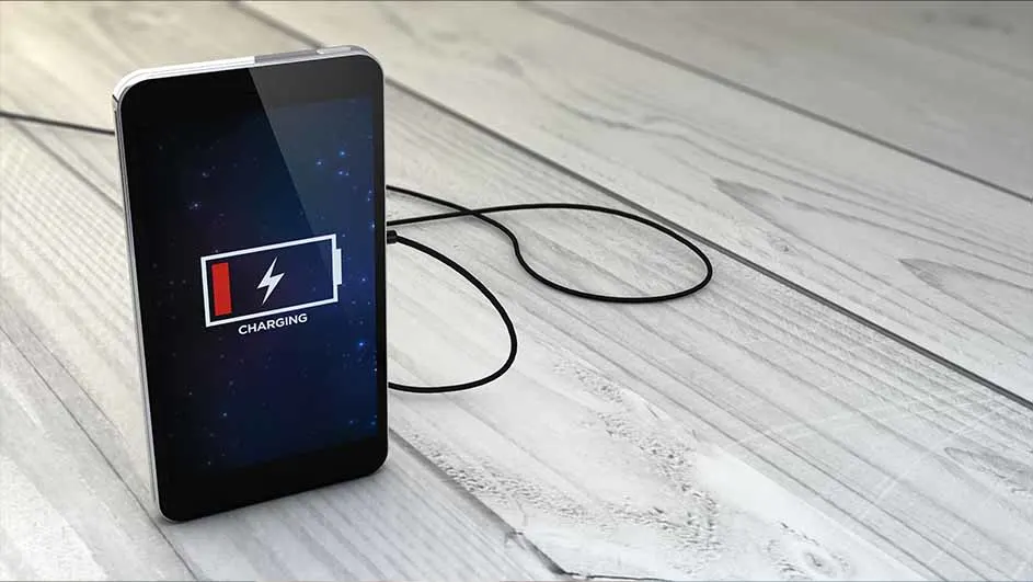Un smartphone noir avec un câble de charge sur une table en bois. L'écran affiche une icône de batterie en charge.