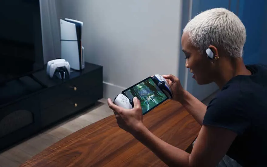 Un joueur aux cheveux gris joue sur une tablette tout en tenant une manette sans fil, assis à une table en bois avec une PlayStation 5 et des accessoires de jeu visibles en arrière-plan.