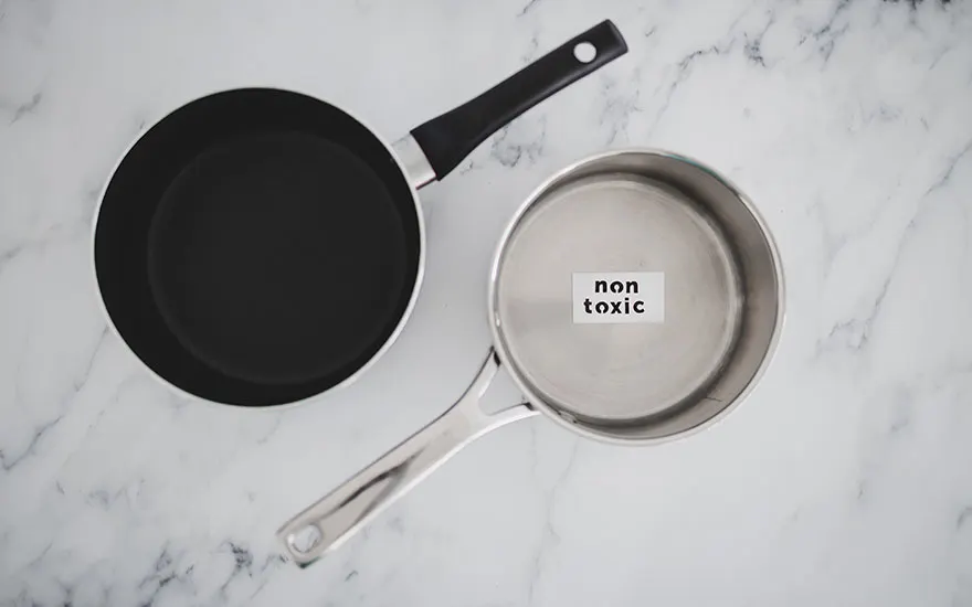 Vue de dessus de deux casseroles sur un plan de travail en marbre. L'une est noire avec une poignée noire, l'autre est argentée avec une étiquette "non toxique" à l'intérieur.