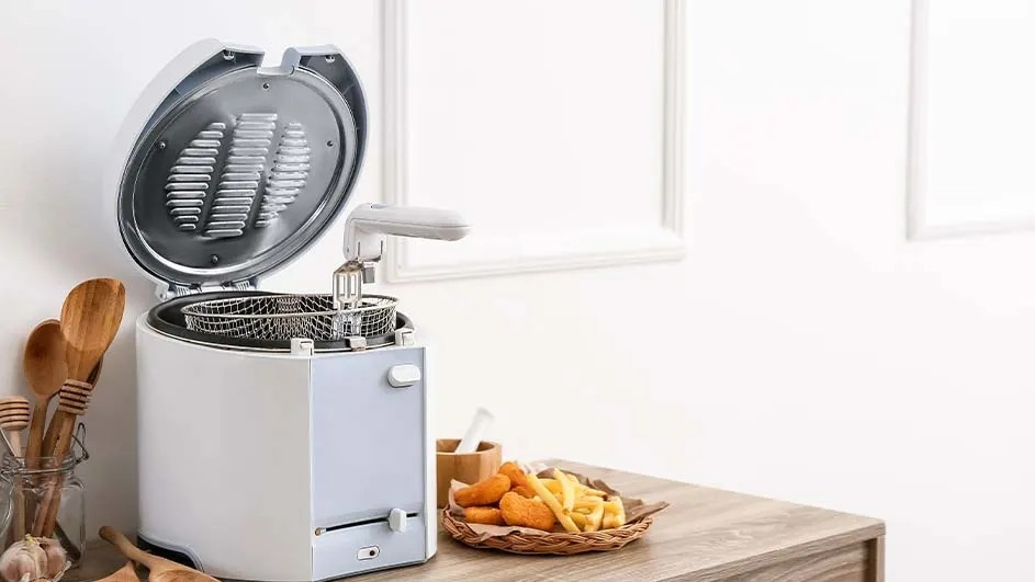 Une friteuse blanche ouverte avec un panier, à côté d'une assiette de frites sur une table en bois devant un mur blanc.