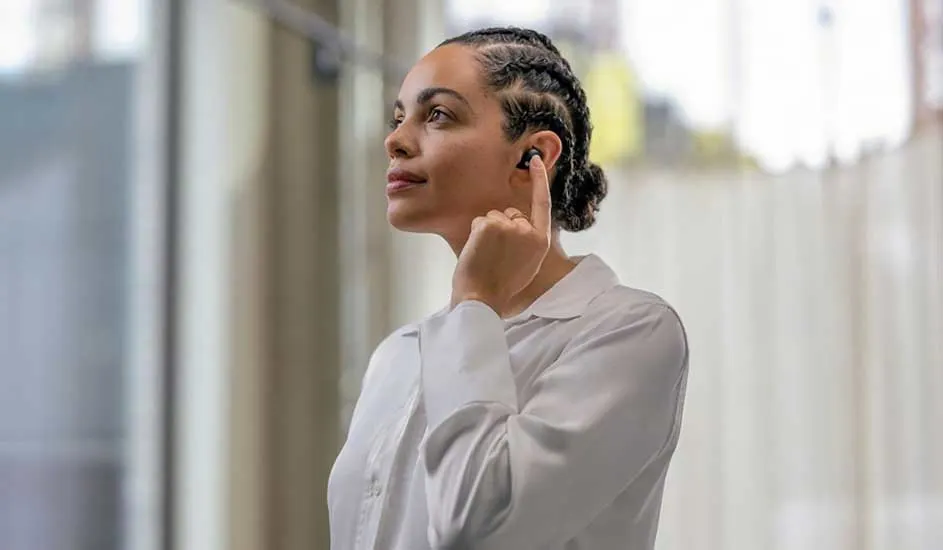 Une personne en chemise claire ajuste un écouteur noir. Rideaux clairs en arrière-plan.