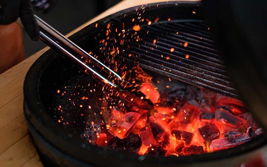 Un barbecue noir rond avec des braises rougesoyantes et des étincelles. Une grille métallique et des pinces sont visibles.