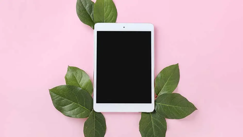 Een witte tablet met een zwart scherm ligt op een roze achtergrond, omringd door groene bladeren.