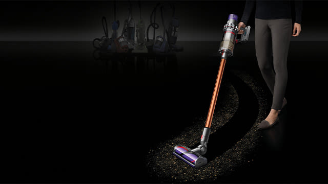 Un aspirateur sans fil orange et argenté sur un sol sombre, avec des meubles flous à l'arrière-plan.