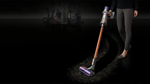 Un aspirateur sans fil orange et argenté sur un sol sombre, avec des meubles flous à l'arrière-plan.
