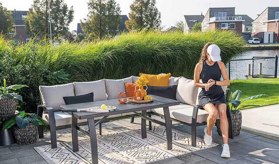 A woman sits on a modern garden bench with cushions beside a table with drinks. Green plants and water in the background. Sunny, peaceful garden with houses and lawn.