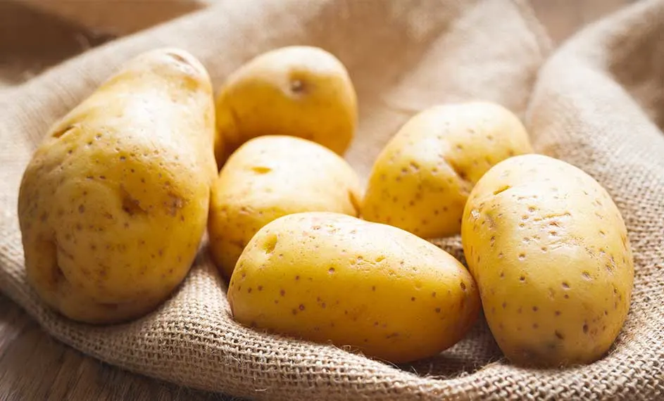 Zes gele aardappelen met kleine bruine vlekjes liggen op een grove, beige doek. De aardappelen hebben verschillende vormen.