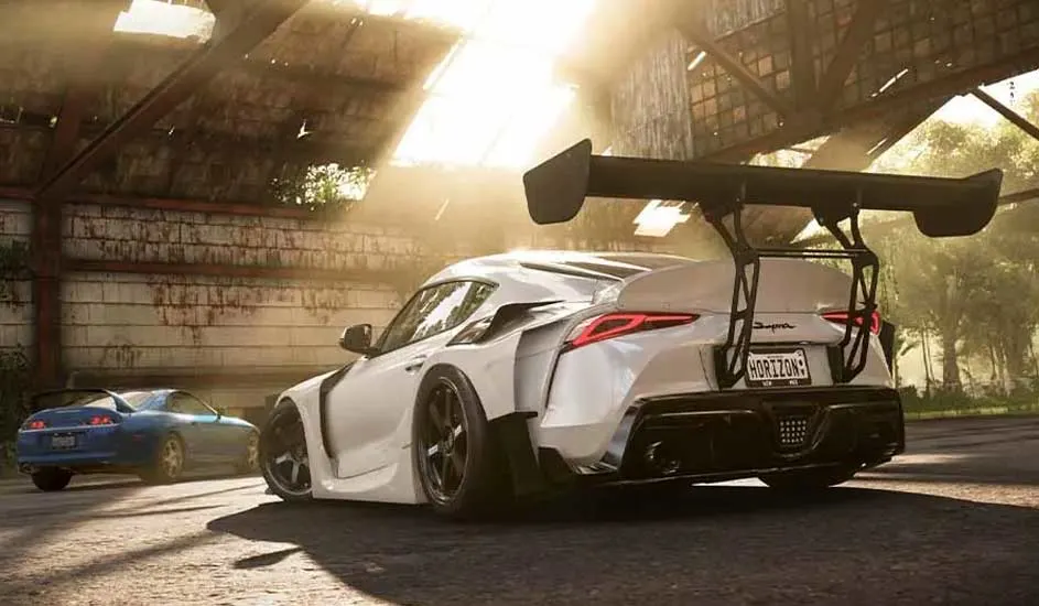 Een witte sportwagen met grote spoiler staat in een garage, een blauwe wagen staat op de achtergrond.