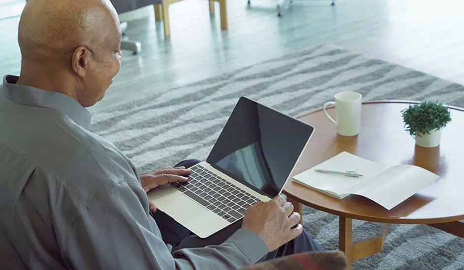 Close-up of a laptop on a lap. A table holds a white mug, a plant, a notebook, and a pen. A gray rug is visible.