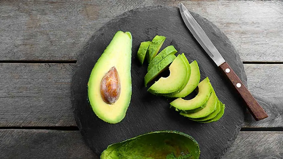 Halve avocado met pit, groene plakjes avocado en een mes op een zwarte plaat, houten tafel.