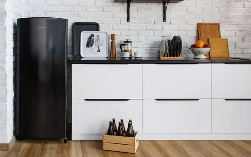 Cuisine avec frigo noir, armoires blanches à dessus noir, et sol en bois. Une caisse de bouteilles est visible.