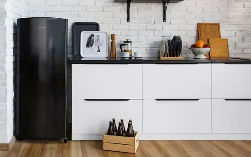 Cuisine avec frigo noir, armoires blanches à dessus noir, et sol en bois. Une caisse de bouteilles est visible.