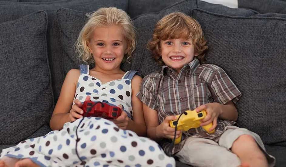 Deux enfants assis sur un canapé gris, tenant des manettes de jeu rouge et jaune.