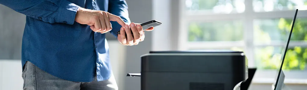 Un homme utilise un smartphone près d'une imprimante noire et d'un écran d'ordinateur, devant une fenêtre lumineuse.