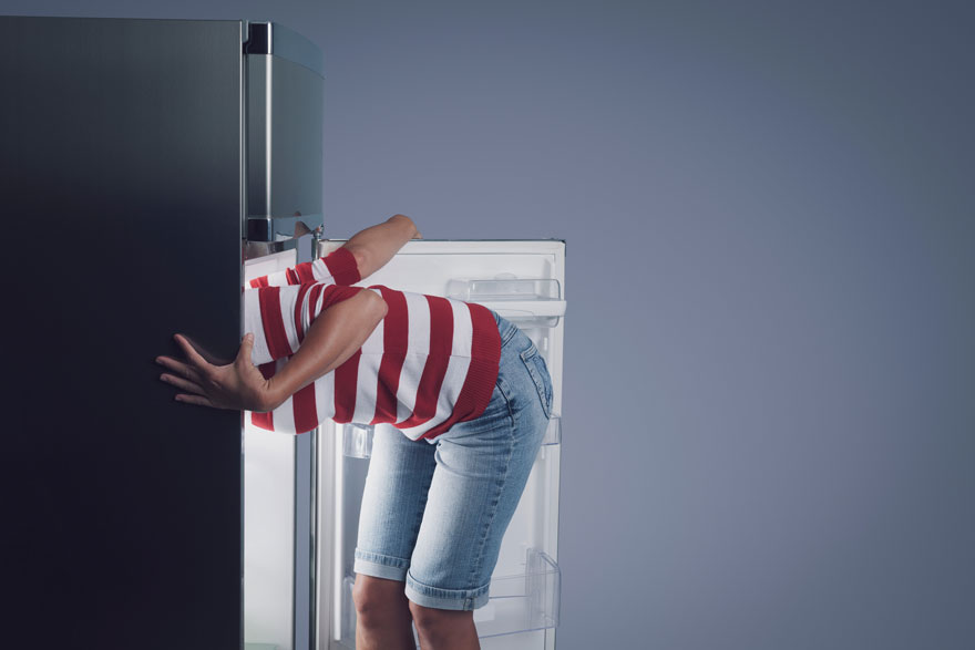 Une personne se penche dans un réfrigérateur noir et blanc ouvert, portant un t-shirt rayé rouge et blanc et un short en jean. Le mur est bleu.