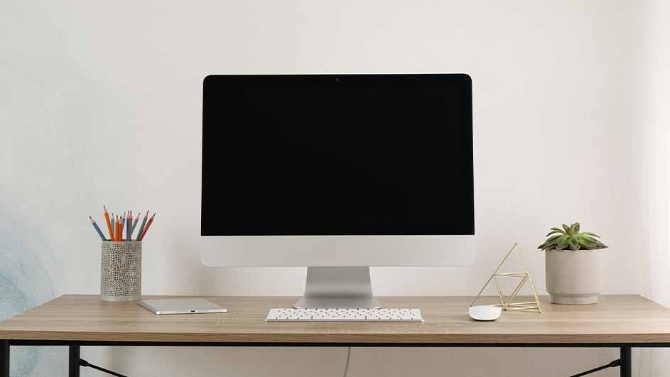 Apple iMac sits centrally on a wooden desk with keyboard, mouse, plants and pen holder; Apple logo visible beneath the screen
