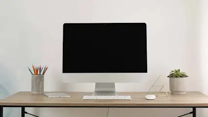 Apple iMac sits centrally on a wooden desk with keyboard, mouse, plants and pen holder; Apple logo visible beneath the screen