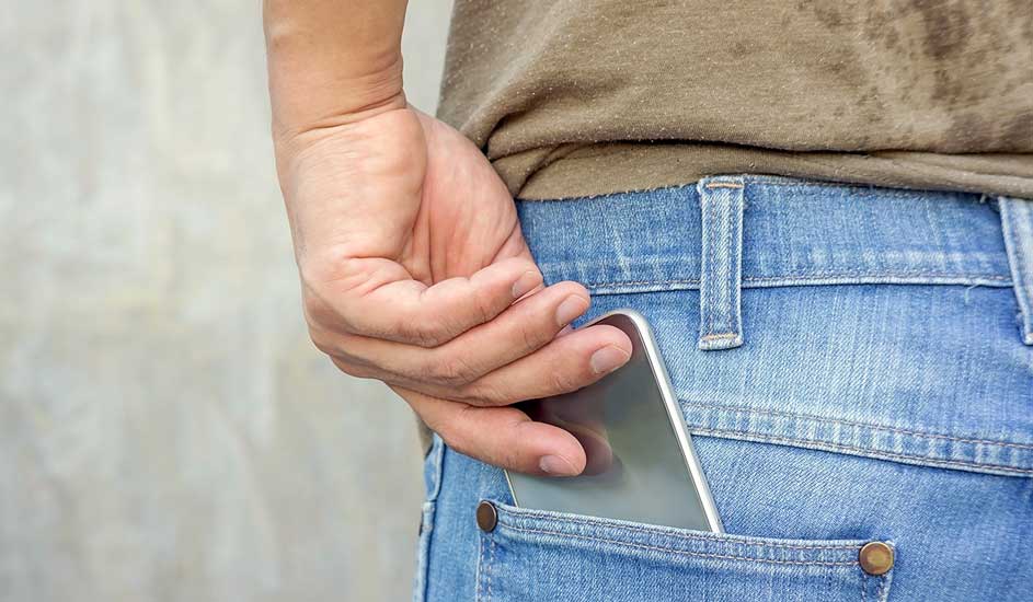 Een zilverkleurige smartphone zit in de achterzak van een blauwe jeans, hand nabij. Grijze achtergrond.