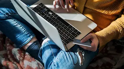 Een open laptop met een extern opslagapparaat aangesloten op een spijkerbroek op een bedsprei.