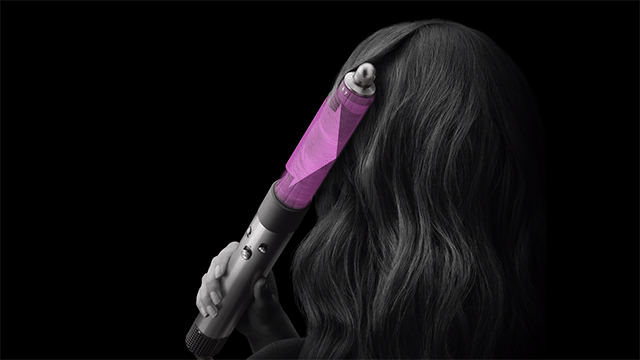 Une personne utilise un fer à boucler violet sur des cheveux ondulés devant un fond noir.