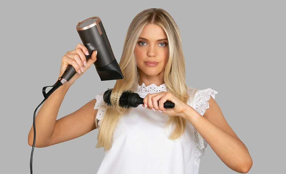Une brosse ronde noire et un sèche-cheveux noir et or coiffent des cheveux blonds sur un fond gris.
