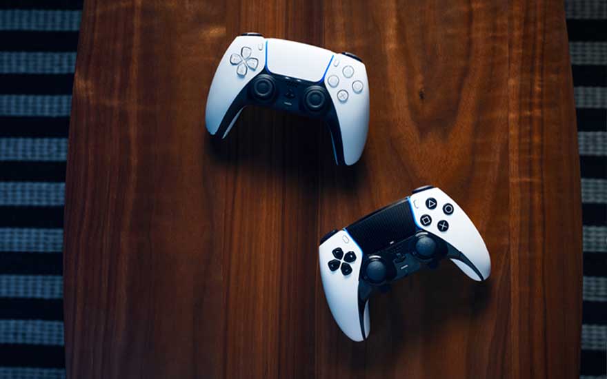 Deux manettes de jeu sont exposées sur une table en bois : une manette PlayStation 5 DualSense blanche et noire en haut et une autre manette en bas, présentant l'équipement gaming moderne élégamment.