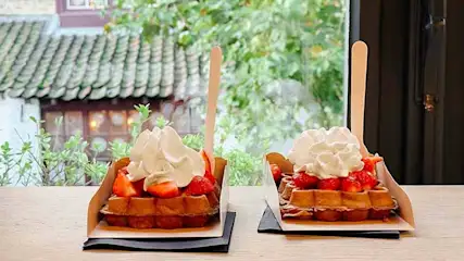 Twee wafels met aardbeien en slagroom staan op een houten tafel, met een houten lepel in elk, voor een raam.