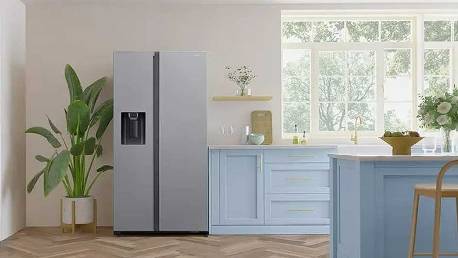 Cuisine lumineuse avec un grand réfrigérateur gris à côté d'une plante en pot et d'armoires bleues sous une fenêtre.