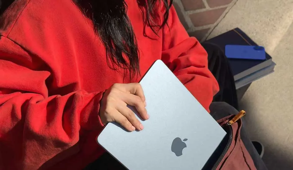 Een grijze laptop met een Apple-logo wordt in een bruine tas gestoken. Op de achtergrond liggen boeken en een blauwe telefoon.
