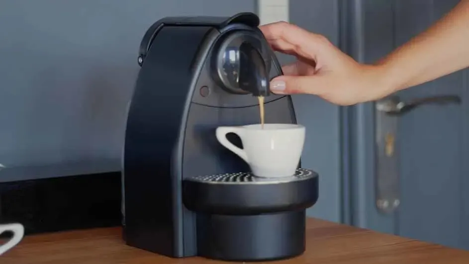 Een zwarte koffiemachine vult een witte kop met koffie op een houten tafel. Een blauwe deur is zichtbaar op de achtergrond.