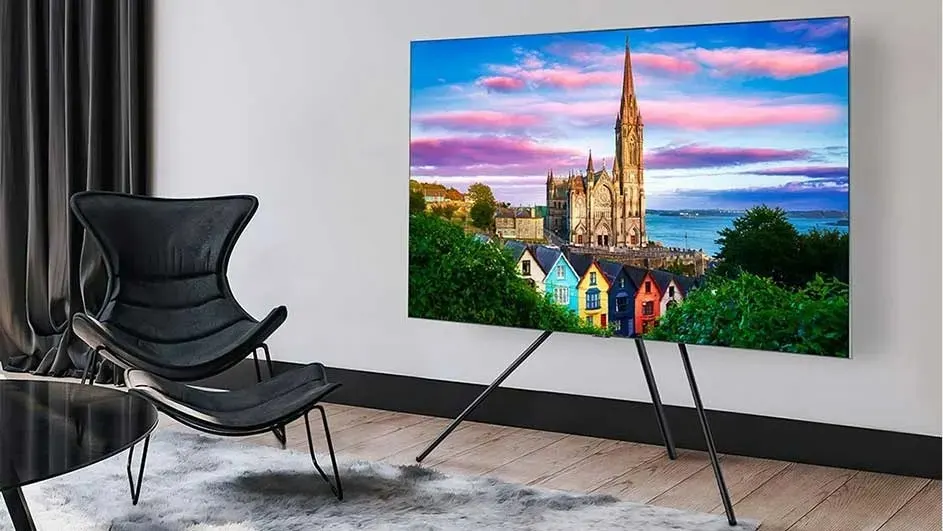 Une TV LG sur trépied affiche une ville colorée et une église, à côté d’un fauteuil design dans un salon moderne et lumineux.