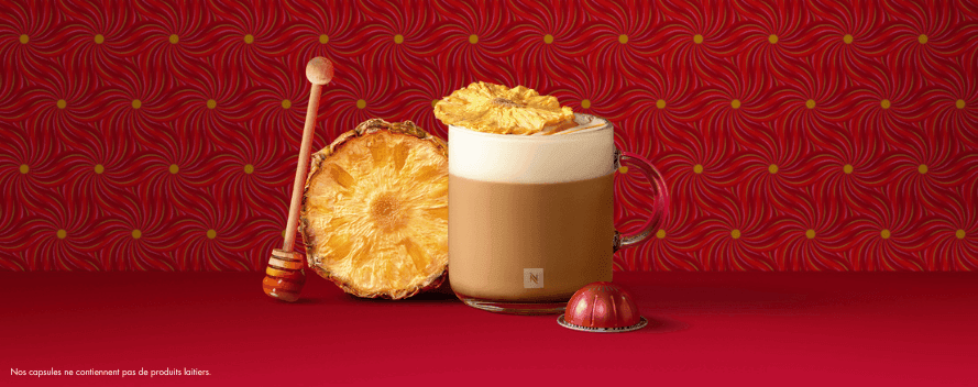 Un café crémeux avec de la mousse de lait, de l’ananas séché et du miel, à côté d’une capsule rouge sur fond rouge à motif floral. Une note en bas indique que les capsules ne contiennent pas d’ingrédients laitiers.