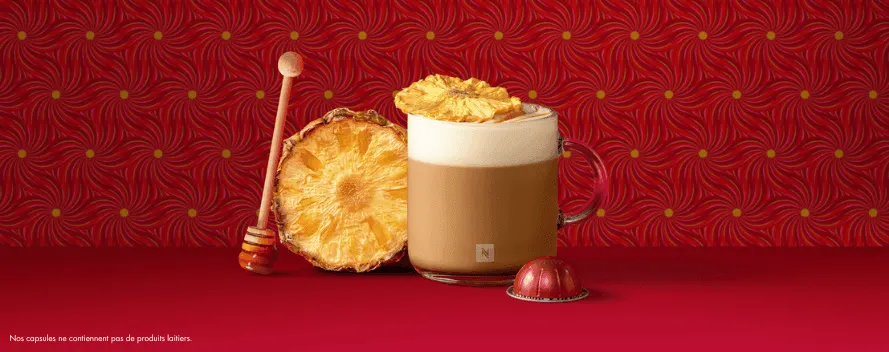 Un café crémeux avec de la mousse de lait, de l’ananas séché et du miel, à côté d’une capsule rouge sur fond rouge à motif floral. Une note en bas indique que les capsules ne contiennent pas d’ingrédients laitiers.