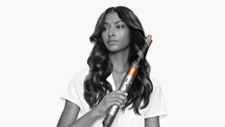 Une femme tient un fer à friser. Elle porte un t-shirt blanc. Ses cheveux noirs sont coiffés en boucles. L'arrière-plan est blanc.