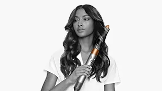 Une femme tient un fer à friser. Elle porte un t-shirt blanc. Ses cheveux noirs sont coiffés en boucles. L'arrière-plan est blanc.
