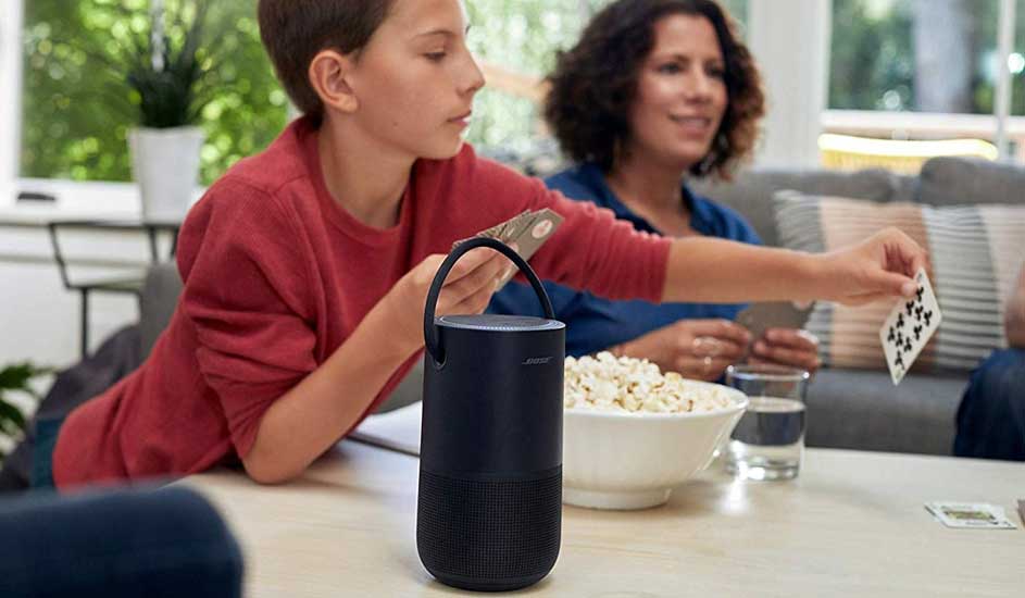 Een zwarte luidspreker staat op een tafel met popcorn, glazen en speelkaarten. Achtergrond: een raam en een bank.