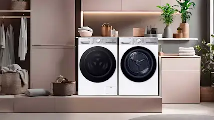 Moderne wasruimte met wasmachine en droger, zachte roze kasten, open garderobe, manden, handdoeken en groene planten.