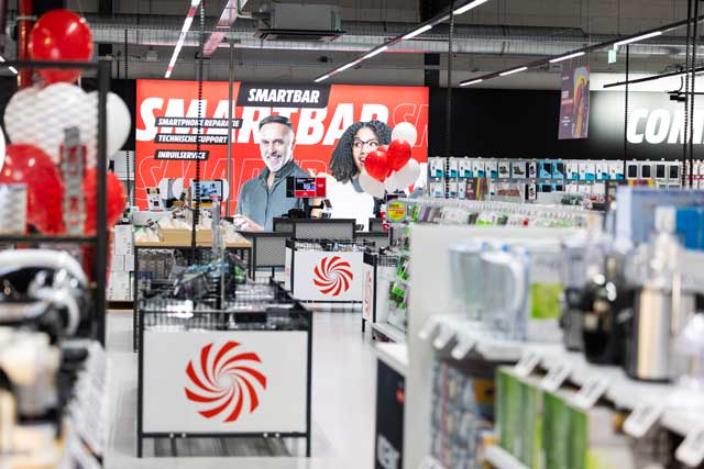 Magasin d’électronique animé avec rayons de gadgets, panneau Smartbar au fond et ballons rouge et blanc au comptoir.