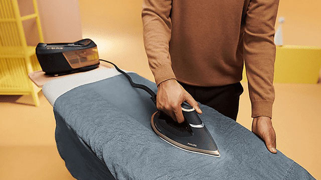 Een stoomstrijkijzer strijkt een blauw kledingstuk op een strijkplank. Op de achtergrond staat een stoomgenerator.