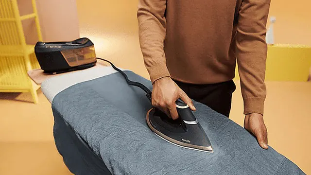 Een stoomstrijkijzer strijkt een blauw kledingstuk op een strijkplank. Op de achtergrond staat een stoomgenerator.