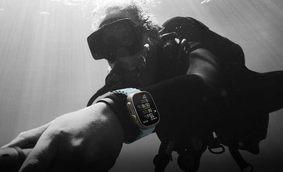 Een zwart-wit foto van een pols met een gouden smartwatch met een blauw bandje onderwater. De achtergrond is wazig.