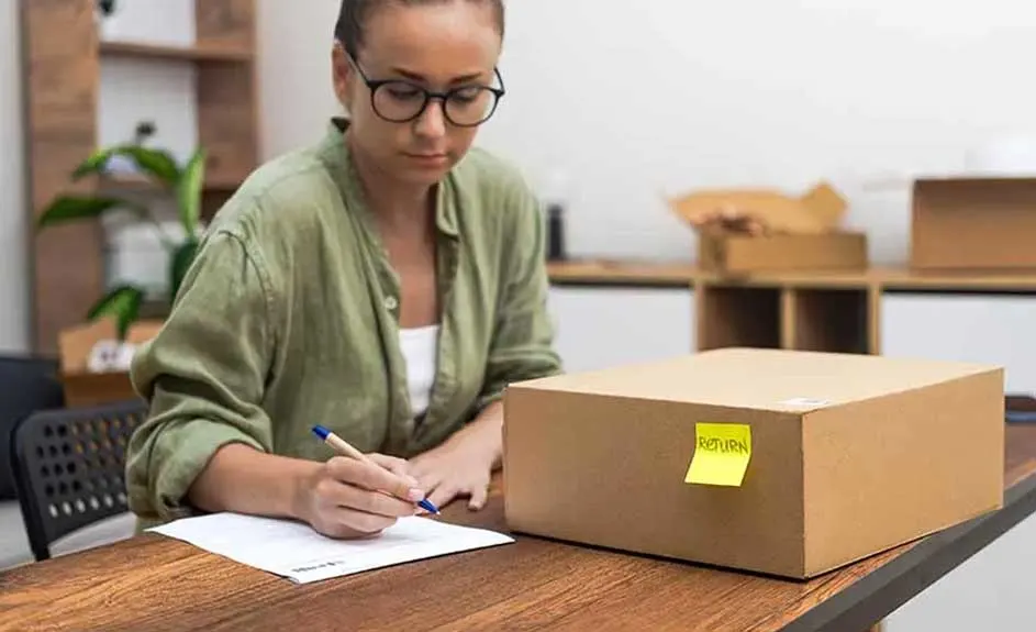 Cardboard box with yellow 'Return' sticker on a wooden desk. A person writes on paper, office items behind.