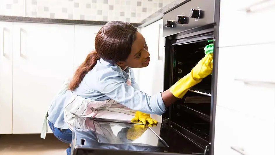 Une cuisine avec un four noir ouvert. Des gants jaunes nettoient l'intérieur. Armoires blanches en arrière-plan.