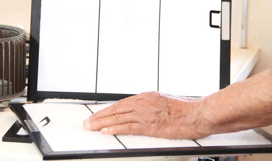 Een hand rust op een wit filter met een zwart frame. Achtergrond toont een ventilator en een groter filter.